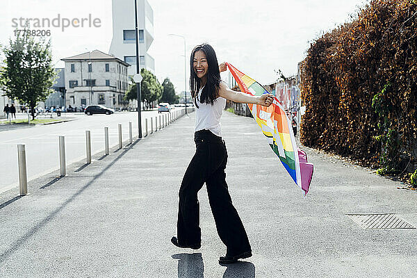 Fröhliche junge Frau mit Regenbogenfahne  die auf der Straße läuft