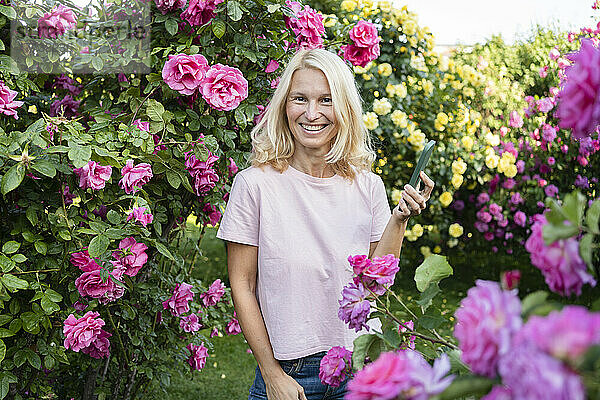 Glückliche Frau mit Mobiltelefon inmitten eines Rosengartens