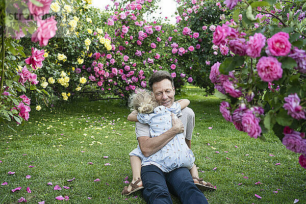 Vater umarmt Tochter  die auf Gras im Rosengarten sitzt