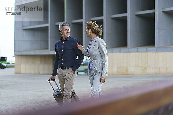 Lächelnder Geschäftsmann mit Koffer  der an einer Geschäftsfrau vor dem Bürogebäude vorbeigeht