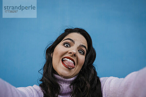 Verspielt zeigt eine glückliche Frau  die ihre Zunge herausstreckt und ein Selfie vor einer blauen Wand macht