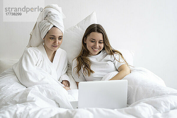 Happy friends reclining on bed and looking at laptop