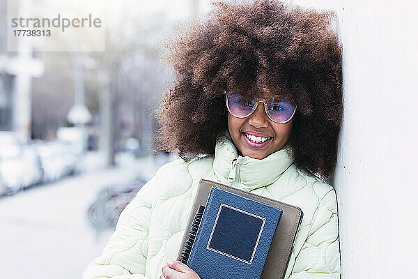 Lächelndes Afro-Mädchen mit Brille und Buch  das an der Wand lehnt