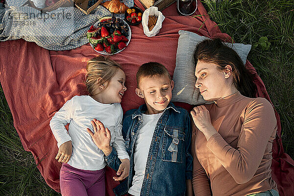 Mutter schaut auf Kinder  die auf einer Picknickdecke im Feld liegen