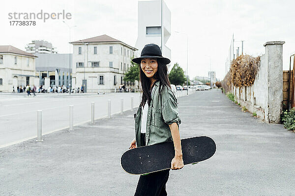 Lächelnde junge Frau mit Skateboard steht auf der Straße