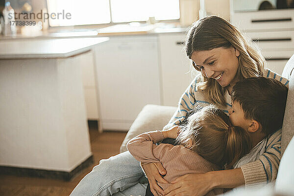 Lächelnde  liebevolle junge Frau umarmt Kinder  die auf dem Sofa sitzen