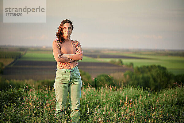 Selbstbewusste junge Frau mit verschränkten Armen steht bei Sonnenuntergang auf dem Feld