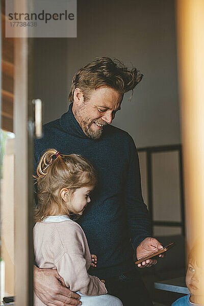 Glücklicher Mann zeigt seiner Tochter durch das Fenster gesehen sein Mobiltelefon