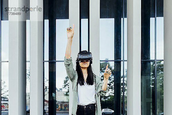 Junge Frau mit Virtual-Reality-Simulator steht mit erhobener Hand vor der Säule