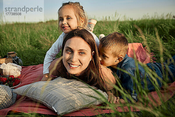 Lächelnde Mutter liegt neben Kindern in Picknickdecke auf dem Feld