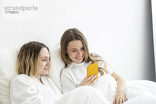 Happy friends using smart phone on bed at home