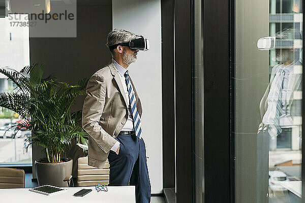 Geschäftsmann mit Virtual-Reality-Simulator steht mit Händen in den Taschen am Fenster im Büro
