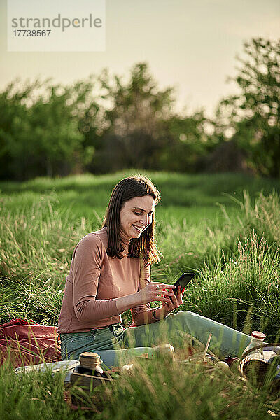 Lächelnde junge Frau benutzt Smartphone beim Picknick im Feld