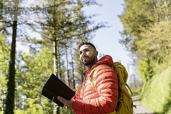 Mann mit Rucksack steht an sonnigem Tag mit Tagebuch im Wald