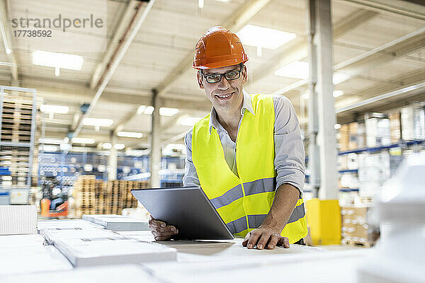 Lächelnder Arbeiter mit Tablet-PC lehnt an Karton im Lager