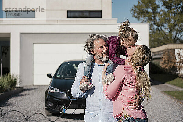 Frau küsst Tochter  die auf der Schulter eines Mannes vor einem Elektroauto sitzt