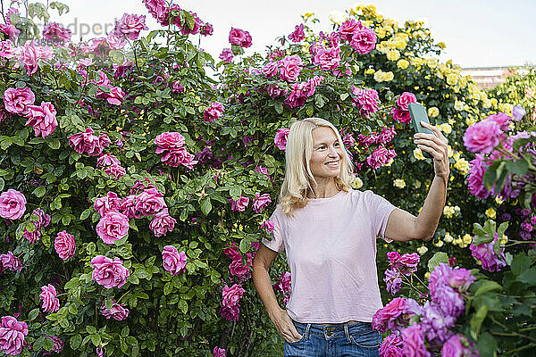 Lächelnde Frau  die mit einer Hand in der Tasche und ihrem Mobiltelefon ein Selfie vor Rosenpflanzen macht