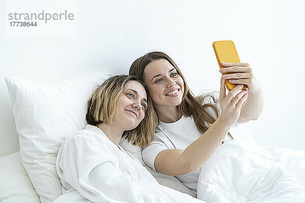 Happy young friends taking selfie through smart phone on bed at home