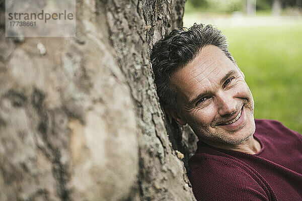 Lächelnder reifer Mann lehnt an Baumstamm im Park