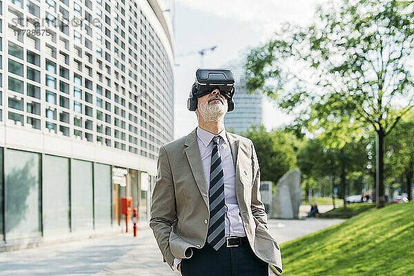 Geschäftsmann mit Händen in den Taschen  der einen Virtual-Reality-Simulator trägt und vor einem Gebäude steht