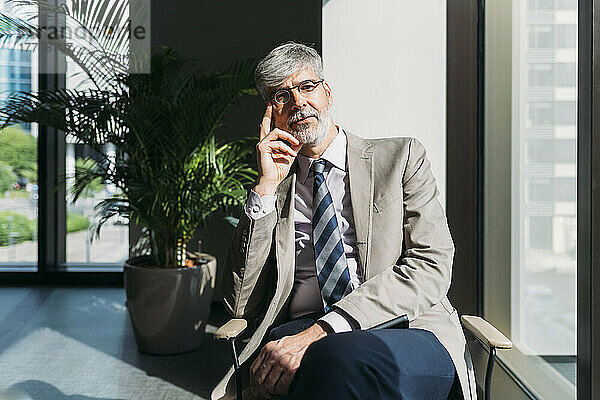 Reifer Geschäftsmann mit Brille sitzt auf einem Stuhl am Fenster im Büro