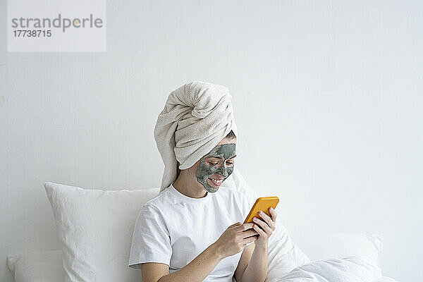 Glückliche Frau mit Gesichtsmaske und Smartphone im Schlafzimmer
