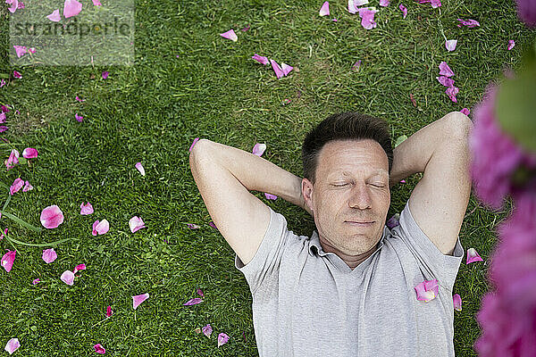 Mann mit geschlossenen Augen liegt auf Gras mit Rosenblättern im Garten