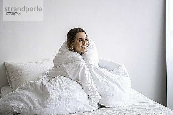 Glückliche Frau  in eine Decke gehüllt  sitzt zu Hause im Bett