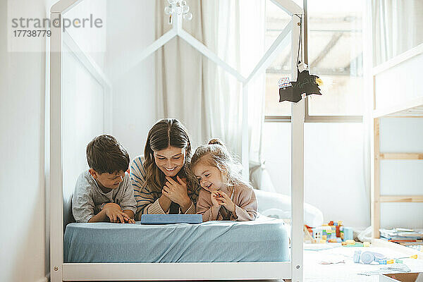 Lächelnde Frau mit Tochter und Sohn  die auf den im Bett liegenden Tablet-PC schauen