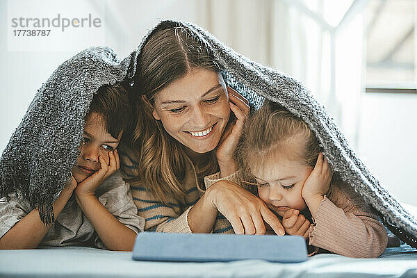 Lächelnde junge Frau und Kinder unter einer Decke  die auf den Tablet-PC schauen  der auf dem Bett liegt