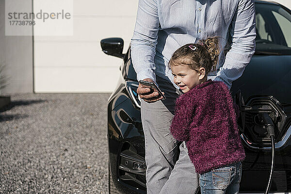 Lächelndes Mädchen  das neben seinem Vater steht und sein Smartphone im Auto hält