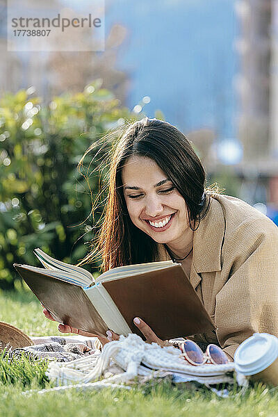 Happy young woman with dark hair reading book lying at park