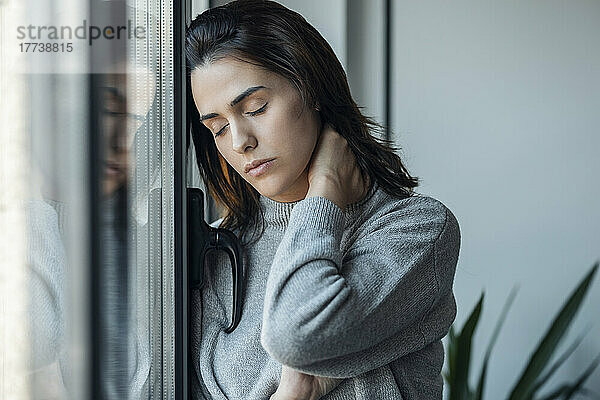Müde Frau mit geschlossenen Augen steht zu Hause am Fenster