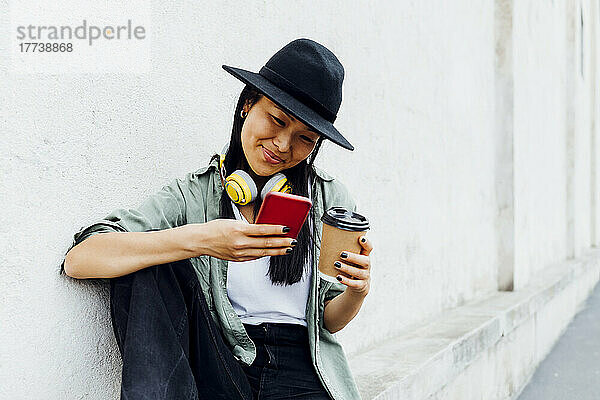 Lächelnde Frau mit wiederverwendbarer Kaffeetasse surft im Internet und nutzt ihr Mobiltelefon  das an der Wand sitzt