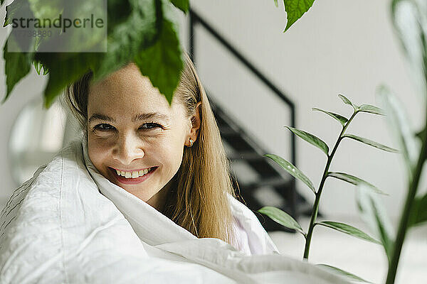 Lächelnde Frau  die zu Hause von Pflanzen in eine Decke gehüllt ist