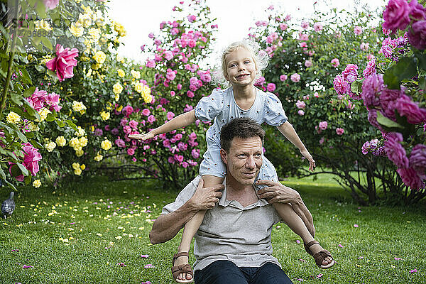 Verspielter Vater trägt Tochter auf Schultern vor blühenden Pflanzen im Garten