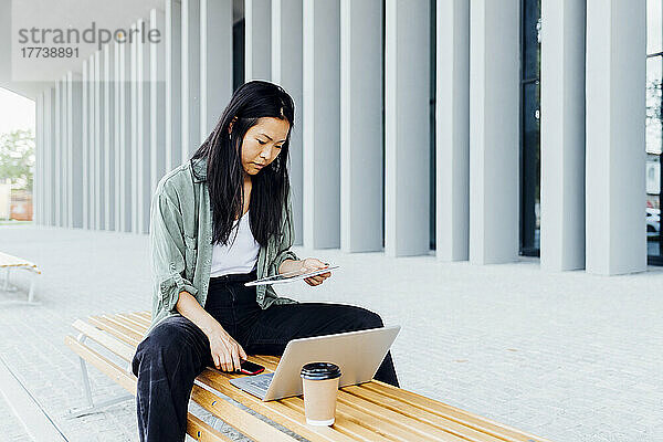 Junger Freiberufler nutzt Tablet-PC  sitzt mit Laptop und wiederverwendbarem Kaffee auf der Bank