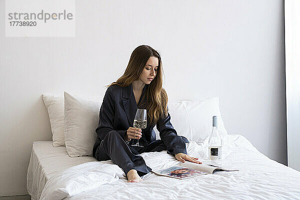 Frau mit Weinglas liest Zeitschrift zu Hause im Bett
