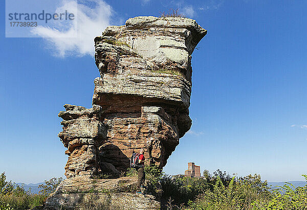 Germany  Rhineland-Palatinate  Senior hiker standing in front of sandstone rock formation in Palatinate Forest with Trifels Castle in distant background