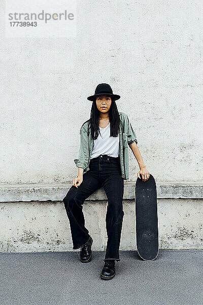 Junge Frau mit Skateboard sitzt an der Wand