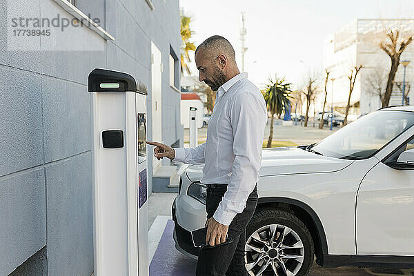 Mann benutzt Automat  der neben dem Auto an der Elektroladestation steht