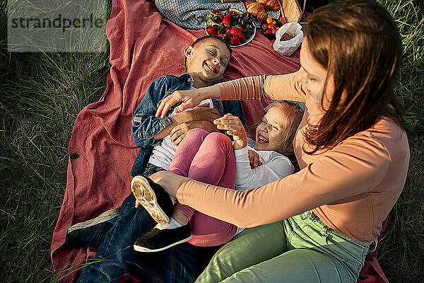 Mutter kitzelt Kinder  die auf Picknickdecke im Feld liegen