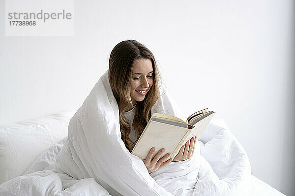 Lächelnde junge Frau  in eine weiße Decke gehüllt  liest zu Hause im Bett ein Buch