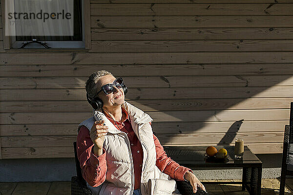 Frau mit Sonnenbrille und Kopfhörer sitzt im Sonnenschein auf der Veranda