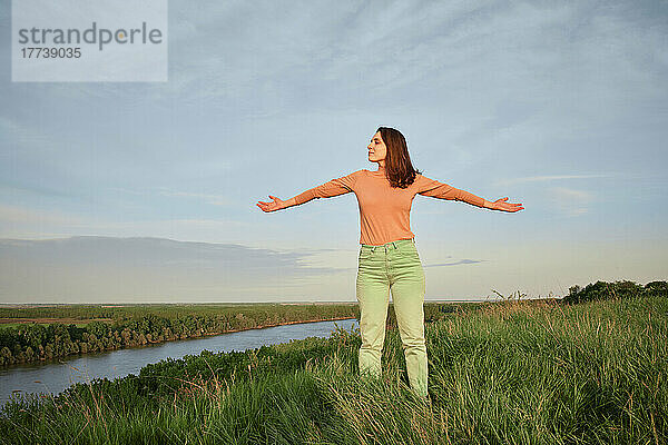 Junge Frau mit ausgestreckten Armen steht auf Gras am Fluss im Feld