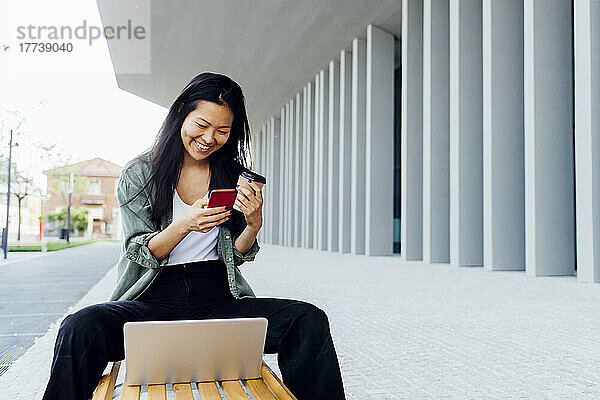 Smiling freelancer holding reusable coffee cup using smart phone sitting on bench