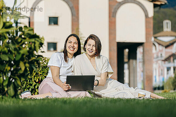 Fröhliche Freundinnen sitzen an sonnigen Tagen mit Laptop im Park
