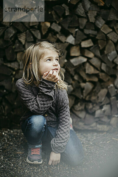 Thoughtful girl with hand on chin kneeling in front of stack of wood