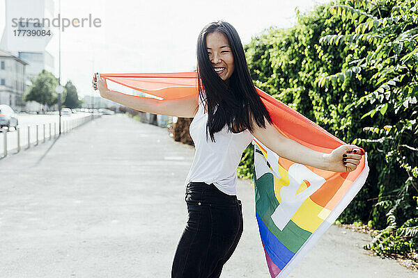 Glückliche Frau mit ausgestreckten Armen  die an einem sonnigen Tag mit einer Regenbogenfahne steht