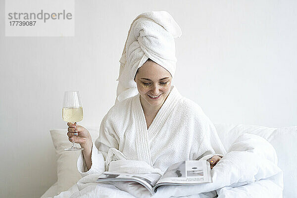 Glückliche Frau mit Weinglas und Zeitschrift sitzt zu Hause im Bett